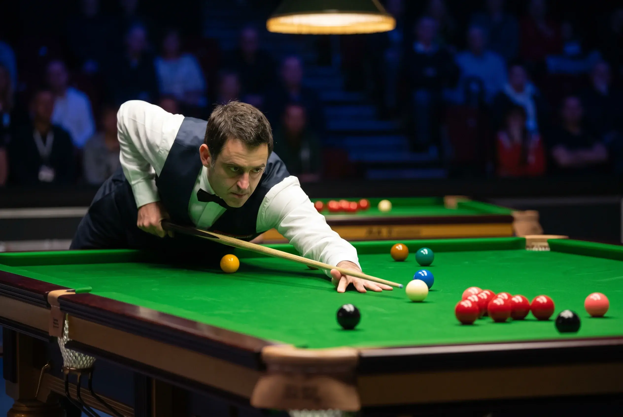 Snookerspieler beugt sich konzentriert über den grünen Snookertisch und zielt auf die schwarze Kugel während eines Live-Matches